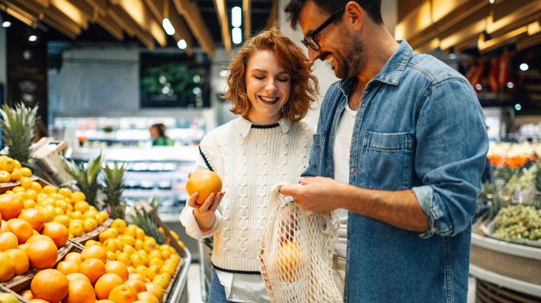 Young smiling couple buying fresh oranges at supermarket, holding reusable mesh bag