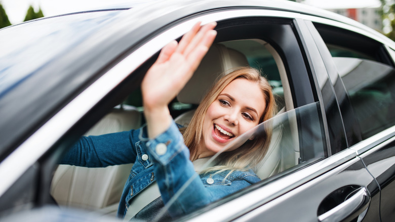 A young woman driver sits in her car and waves toward someone