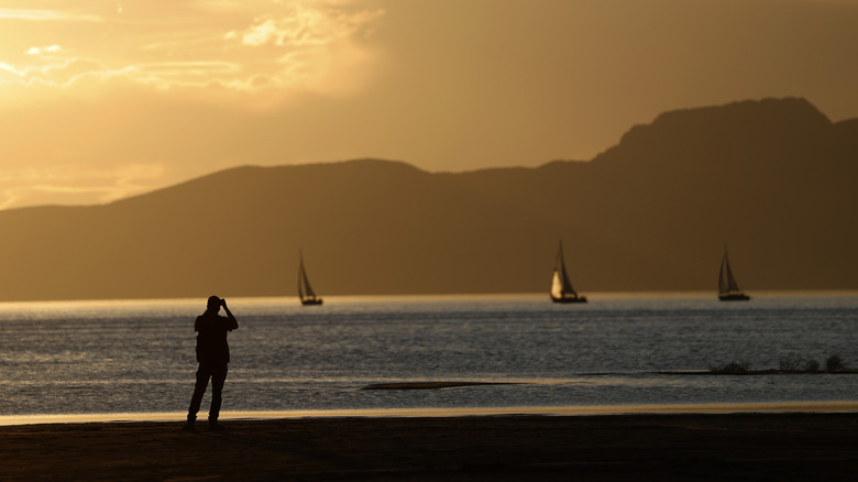 A visitor watches the sunset from the banks of the Great Salt Lake with sailboats on the water.