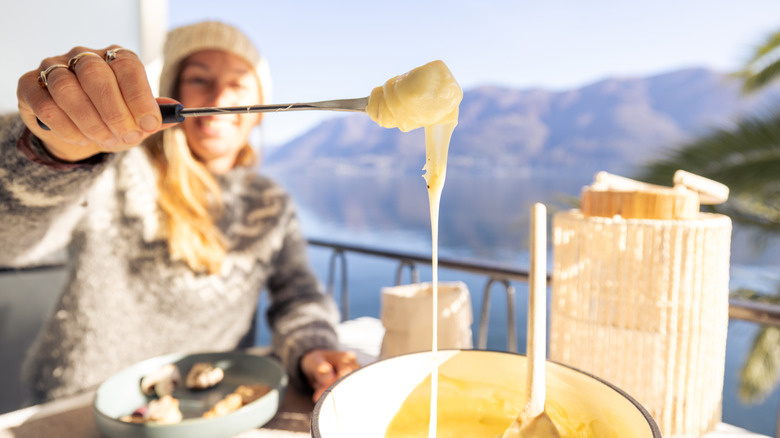 A smiling woman eating cheese fondue with a lake and mountains in the background.