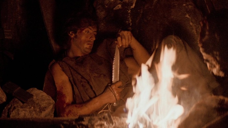 Silvester Stallone looks at knife while sitting in front of fire in "First Blood"