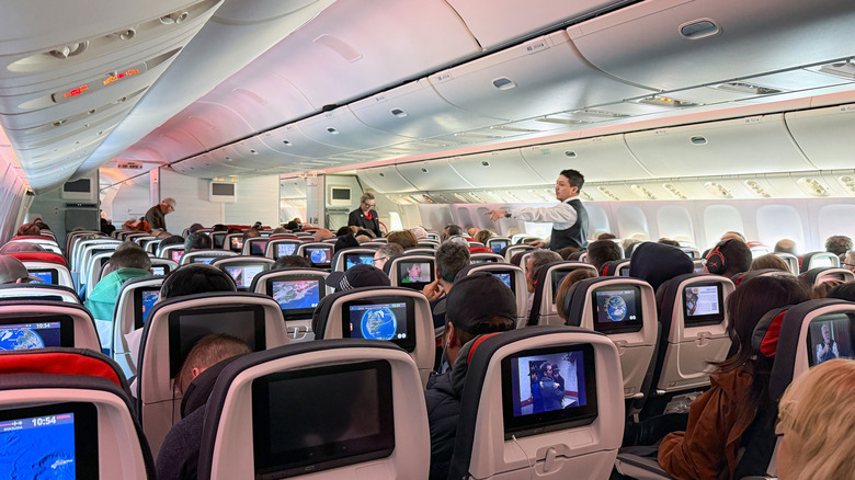 Cabin of an airplane, with a flight attendant standing and passengers watching screens