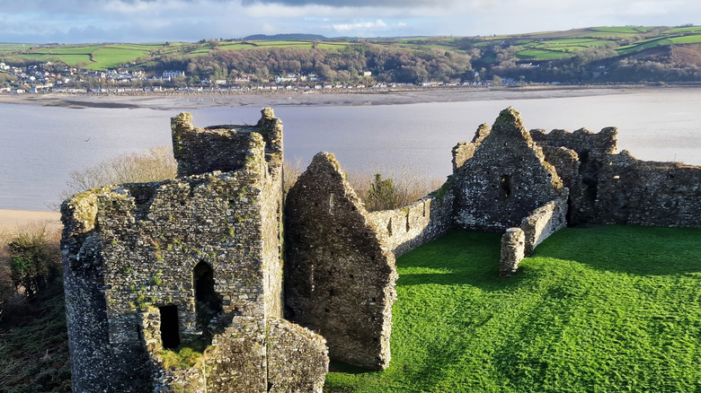 The ruins of Llansteffan Castle in Wales set amongst thick grass overlooking water, buildings, and greenery in the distance