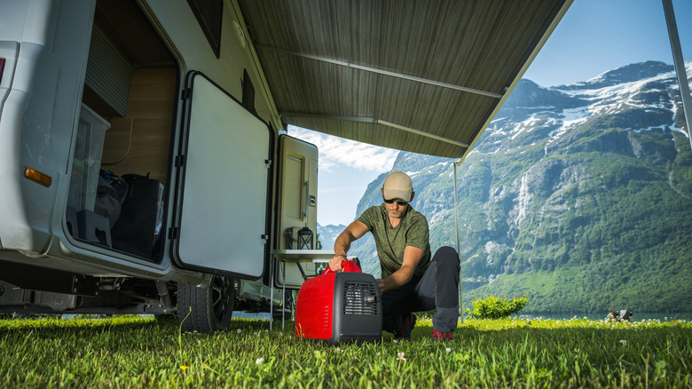 Man crouched beside a red generator next to an RV, with a snow-capped tree-covered mountain in the background