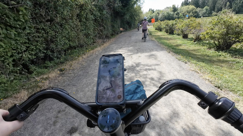 A point-of-view action shot shows a phone attached to bicycle handlebars and other riders pedaling ahead on a gravel trail