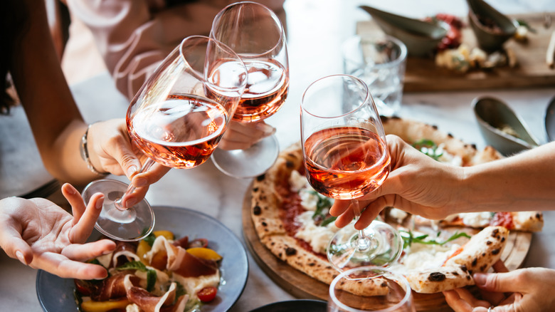 Three people toast glasses of rosé wine over pizza.