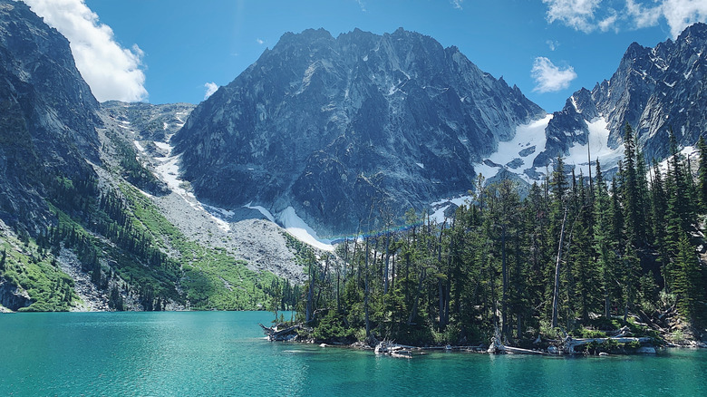Rocky peaks dotted with snow sit high above a crystal blue lake in a glacial valley with a peninsula of pine trees