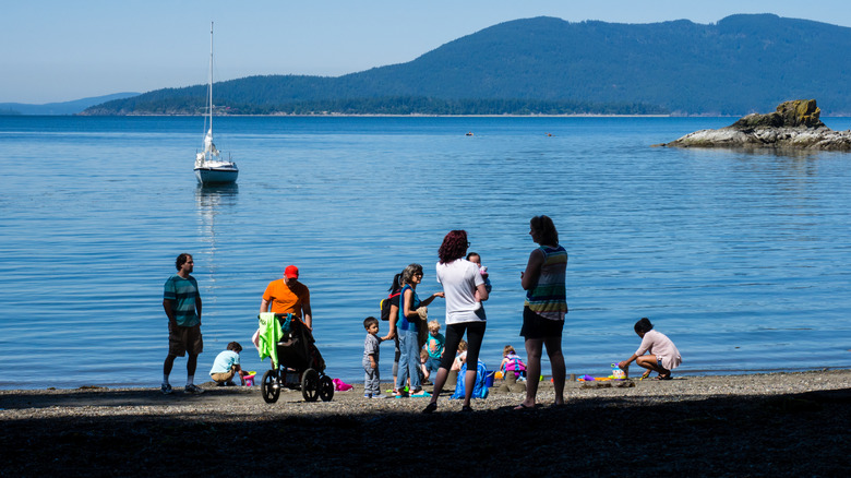 People gather on a pebbled beach on a sunny day as mountains and a sailboat linger in the distance in Larrabee State Park, Washington State