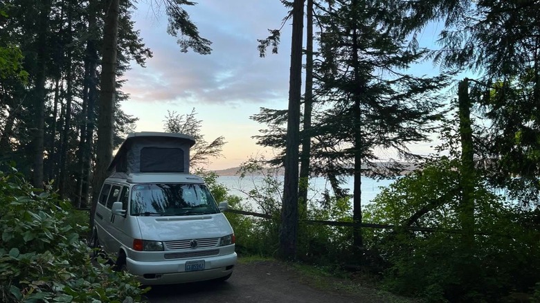 A white camper van parked beneath towering evergreens overlooking the calm water at Sequim Bay State Park, in Washington.
