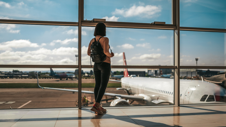 woman staring out airport window at airplane, holding backpack straps