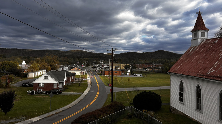 Town of Paw Paw, with a red-roofed white church in the foreground and a two-lane road running past yards and buildings, with treed mountains in the background under a cloudy sky