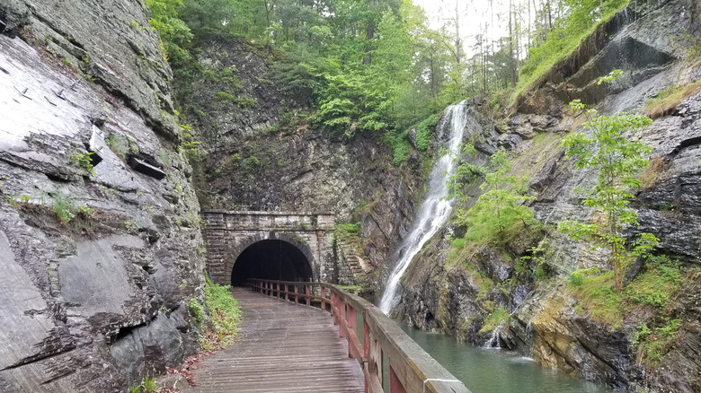 Exit from the Paw Paw Tunnel, with a wooden walkway alongside a canal passing through a stone archway in the mountain, with a waterfall running over the cliff to the right and green trees growing above