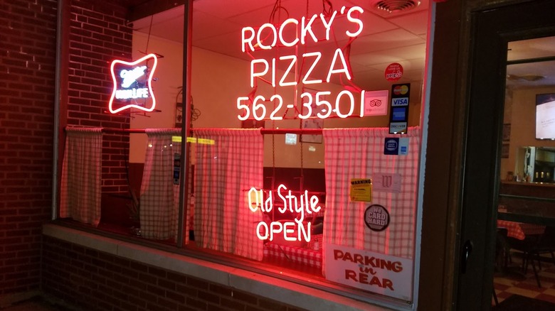 The exterior of Rocky's Pizza, showing a neon sign and checkered curtains