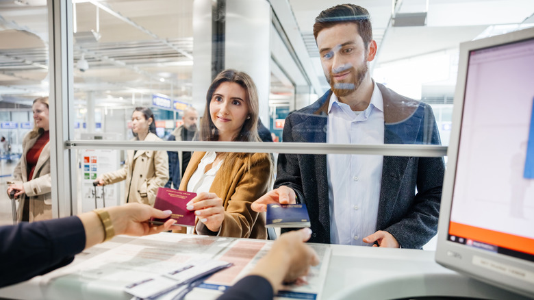A man and woman hand over a red and blue passport to an immigration officer in a glass booth