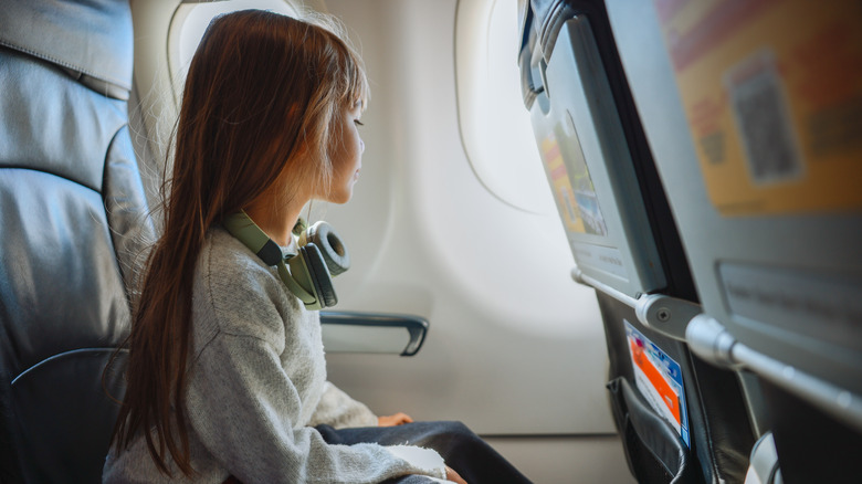 A girl with headphones around her neck sitting on a plane looking out the window.