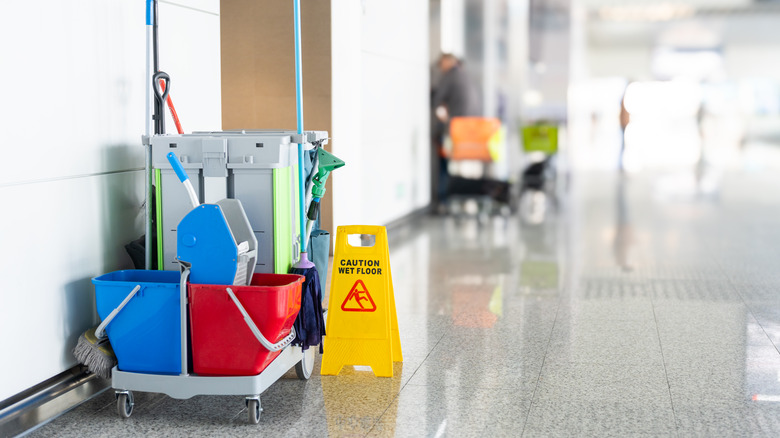 A janitor's cleaning supplies sitting outside a bathroom in an airport terminal with shiny floors.
