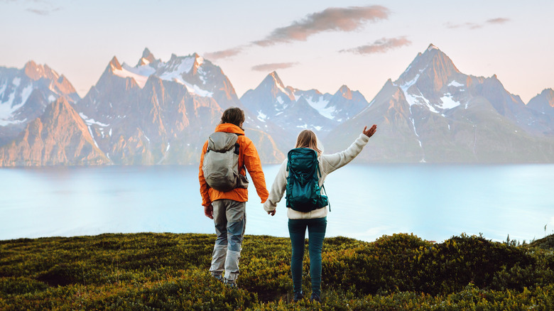 A hiking couple in outdoor gear wearing backpacks are holding hands with their backs to the camera, with mountains in the distance.