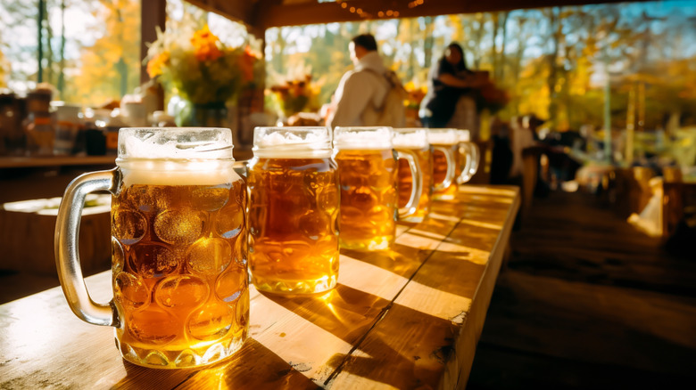 Five steins of beer lined up on a wooden table at an outdoor beer garden.