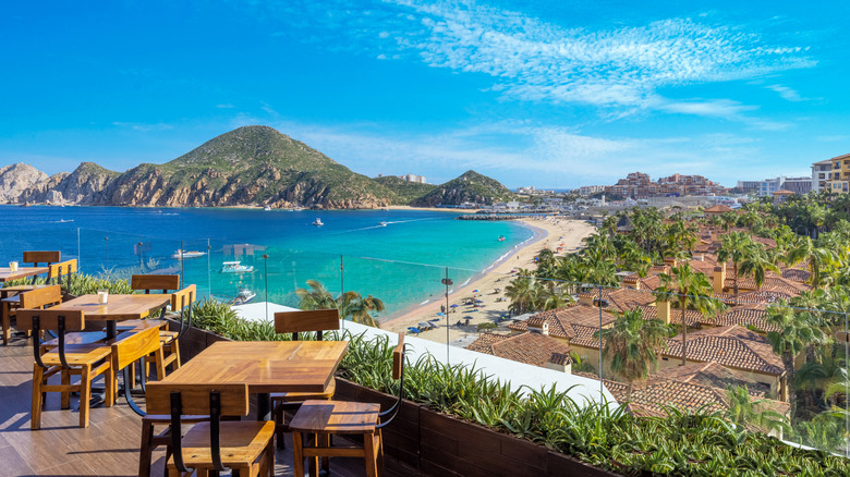 View from balcony with tables and chairs overlooking houses, beach, vibrant water, and hills