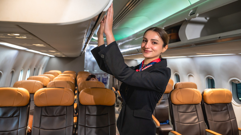A smiling flight attendant closing an airplane's overhead bin.