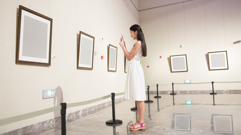 A girl stands in front of blank framed canvases hung on a gallery wall and takes a picture with her phone