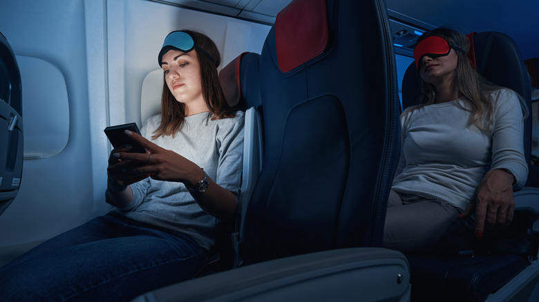 Dark airplane cabin with person on phone with eye mask on head while person behind sleeps