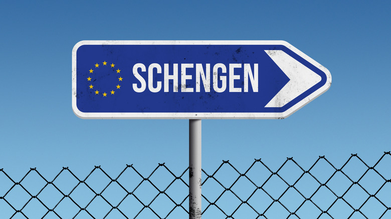 Blue Schengen sign with European flag pointing right against blue sky and fence
