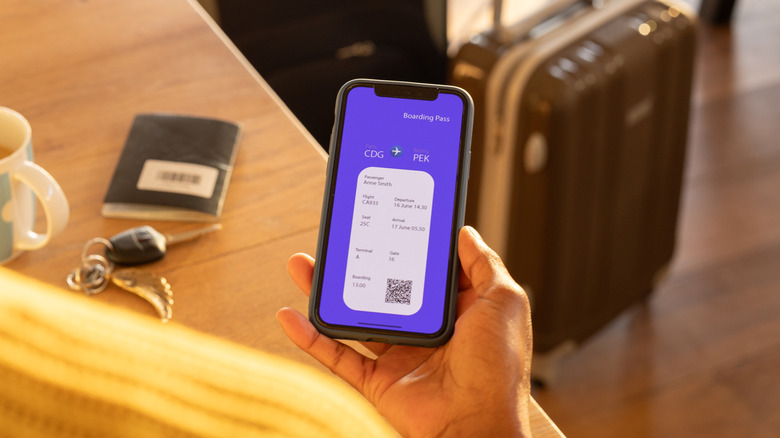 Female traveler with luggage checking her digital boarding pass while at a table with a cup of coffee, a passport, and keys on top of it.