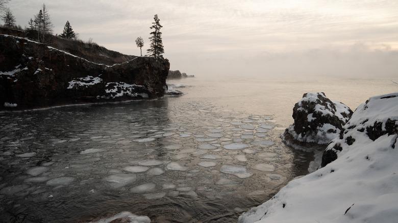 Lake Superior in winter, with ice chunks on the lake surface, a tree-and-snow dotted cliff to the left, and snow-covered land in the foreground, under a gray sky