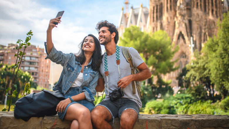 A male and female traveler sit on a wall in a public park near Sagrada Familia in Barcelona, taking a selfie on a bright summer day