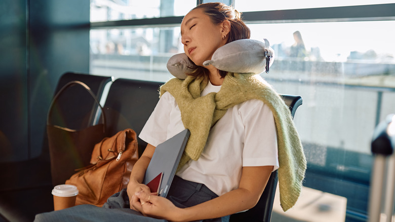 A young Asian woman sleeps using a neck pillow at the gate of her flight in an unidentified airport, with her bags beside her and holding her passport and laptop against her body