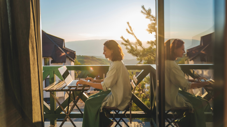 Young woman sitting on a balcony with a view of mountains and sunset, working using a laptop