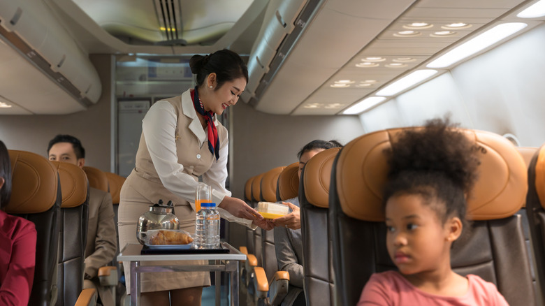 A flight attendant serves a meal to a passenger from a cart stocked with bottled water and a coffee urn