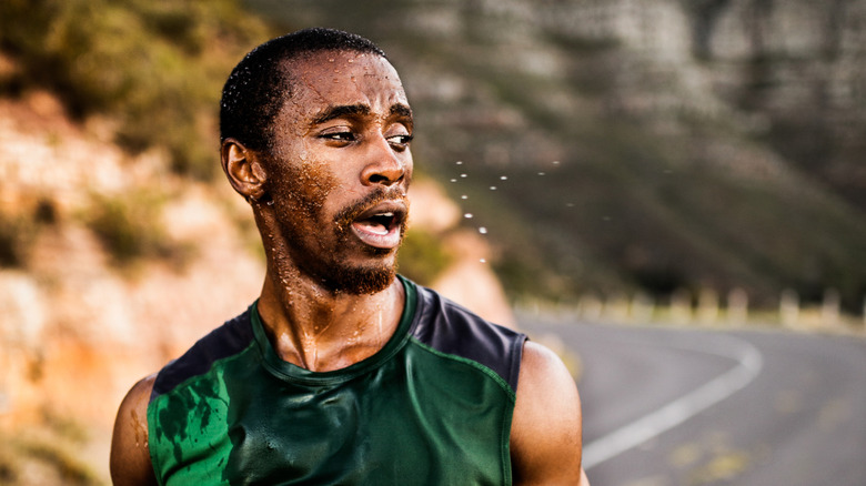 A hot an sweaty man walking down a road with his tank top drenched in sweat.
