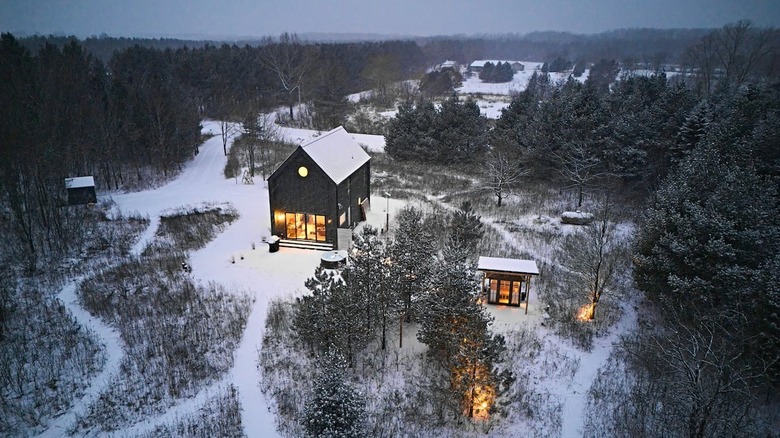 A large black wood-clad home stands in a snowy forest landscape at dusk. There is a small outhouse a few feet away.