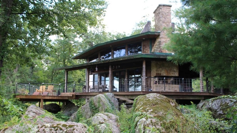 A rustic cabin wiht a chimney and an elevated outdoor deck balanced on large rocks. The house is surrounded by lush trees.