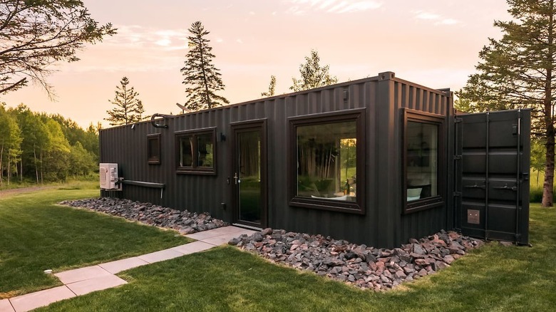 A black shipping container with a door and windows sits on a manicured lawn. There is a forest in the background.