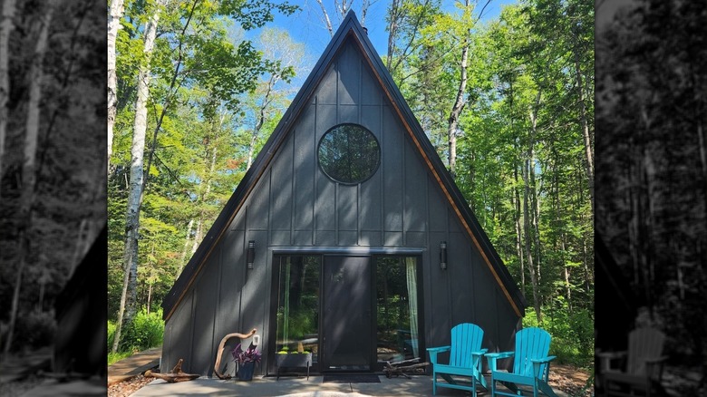 A black A-frame cabin with porthole windows and sliding glass door against blue skies. Two blue-green patio chairs in the foreground.