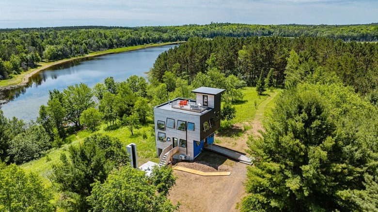 A contemporary, multi-level modern home with upper and lower decks amid greenery and a river behind it.
