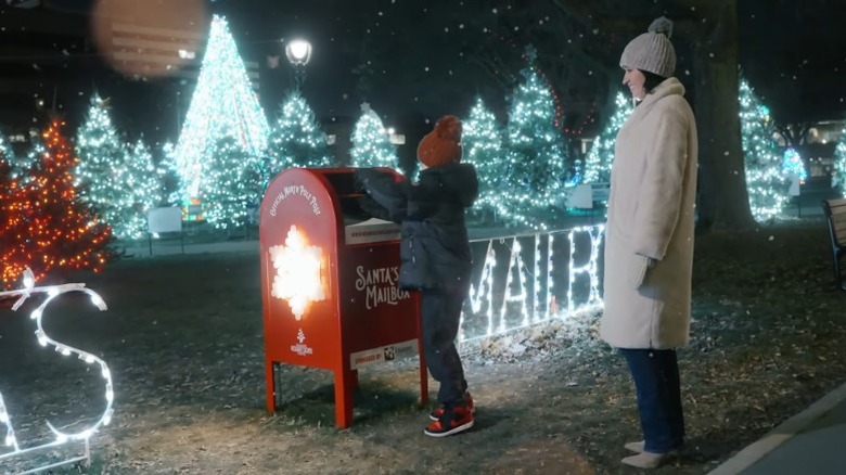 A mother watches her child post a letter to Santa surrounded by Christmas trees and lights