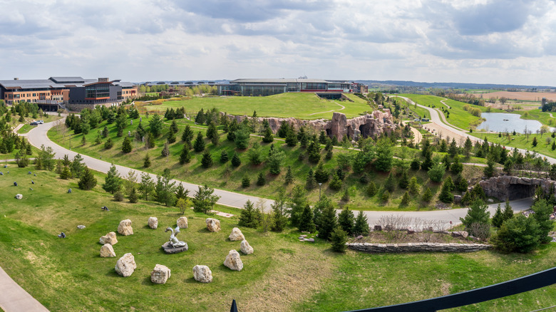 Bird's eye view of tree-dotted hills, a lake, and building. Stone sculptures in the foreground.