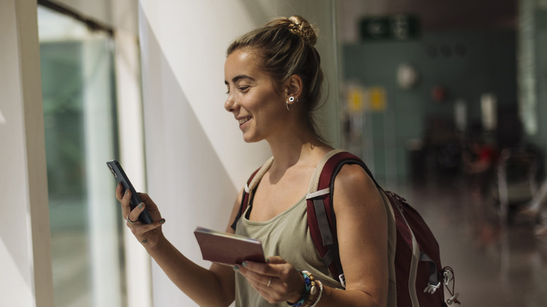 A person looking at her phone, holding a book, and wearing a backpack
