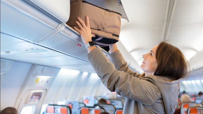 A woman puts a suitcase into a modern airline overhead luggage bin.