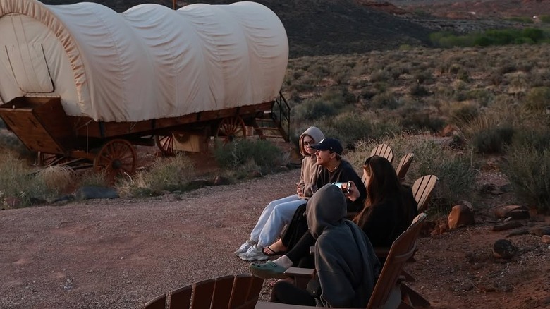 Group of people sitting around a fire pit near a covered wagon glamping setup at Zion Wildflower Resort, near Zion National Park in Utah