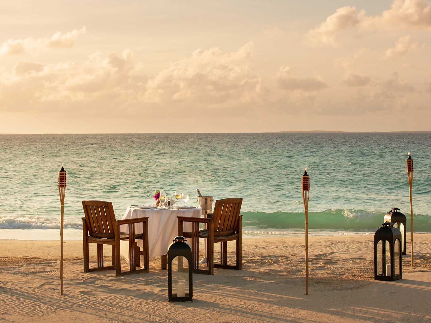 Tables, chairs, and torches set up along the shoreline of a beach.