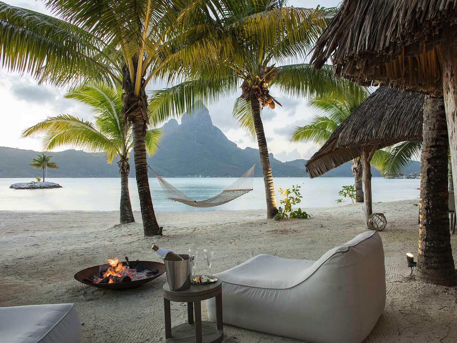 Palm trees, lounge furniture, and a hammock on the beach of Bora Bora. The ocean and a mountainous island is in the distance.