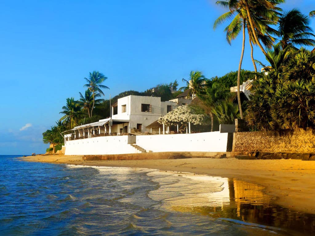 A white building on a beach front surrounded by luscious palms.