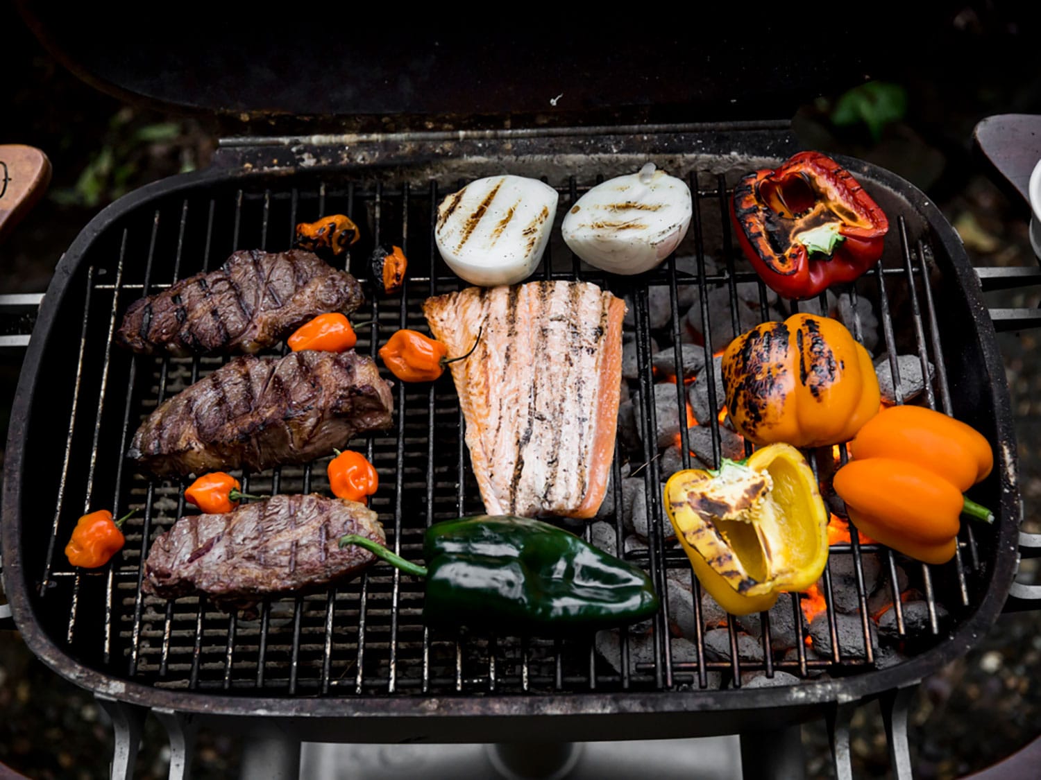 An open PK Grills PK360 grill and smoker being used to cook multiple steaks, fish, and peppers.
