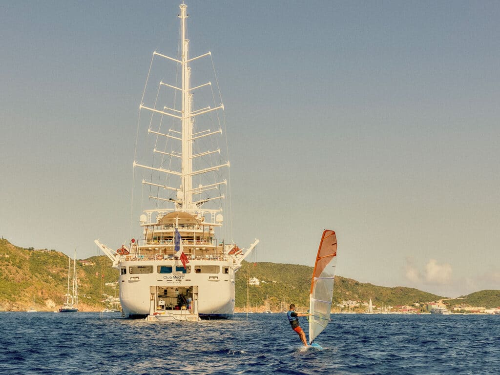 The rear view of the Club Med 2 luxury sailing yacht with a windsurfer following.