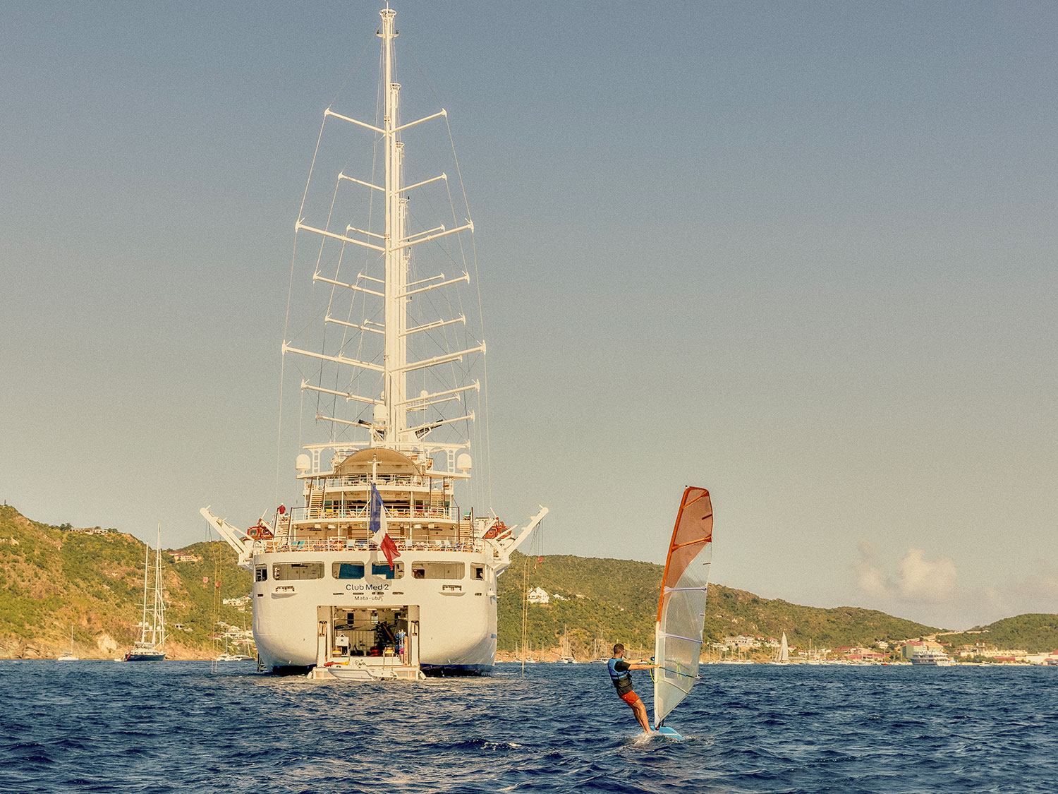 The rear view of the Club Med 2 luxury sailing yacht with a windsurfer following.